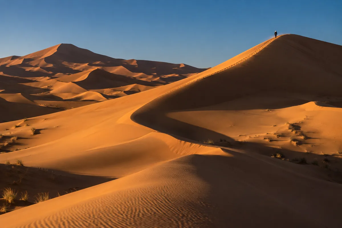 Silhouette d’un marcheur vu de loin sur une crête de dune, minuscule face à l’immensité du sable